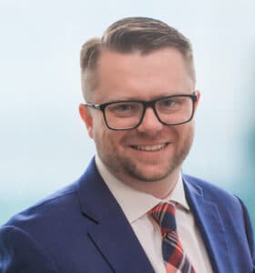 A man with short hair and glasses, wearing a blue suit, white shirt, and a plaid tie, smiles at the camera. The background is softly blurred, suggesting an indoor setting.
