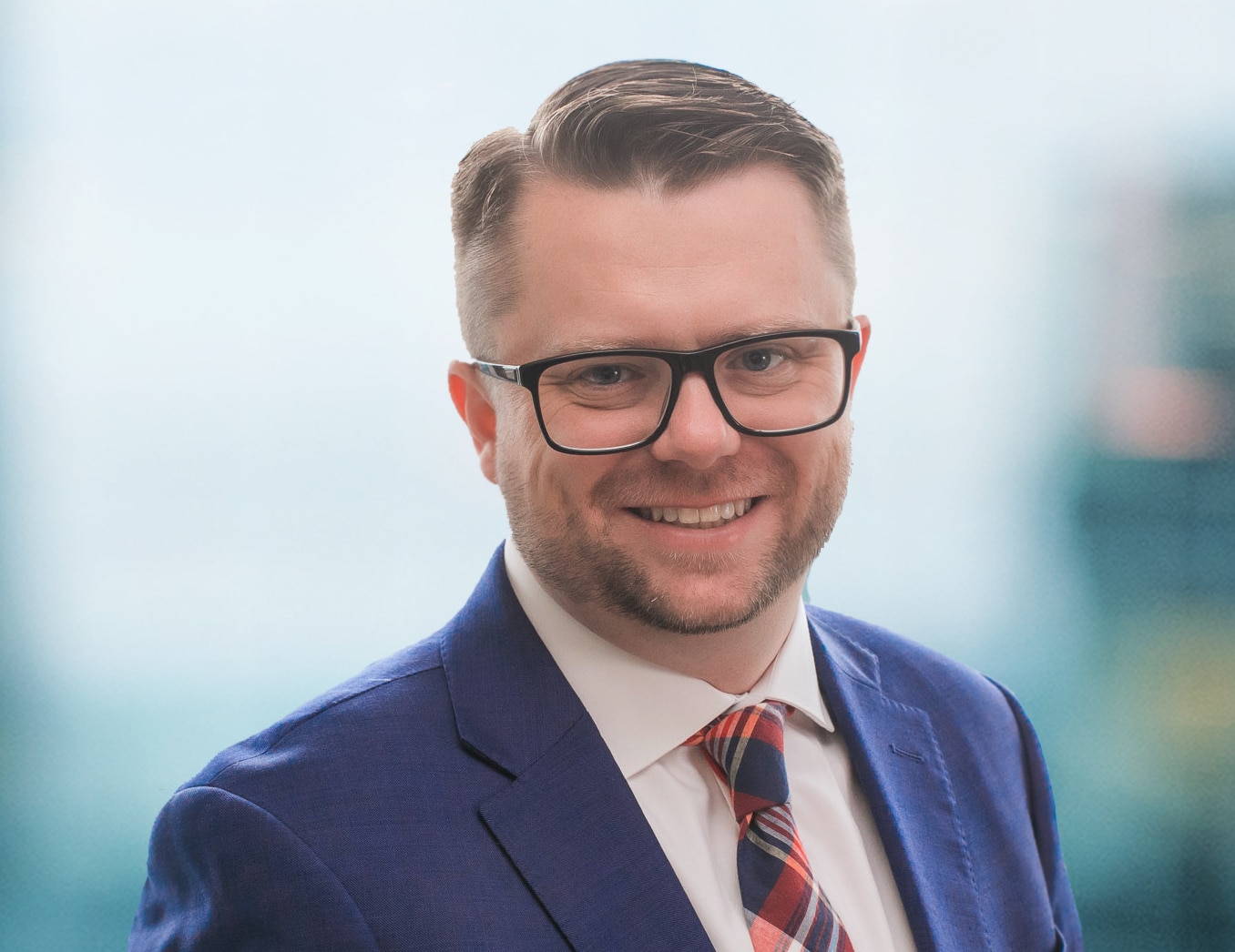 A man with short hair and glasses, wearing a blue suit, white shirt, and a plaid tie, smiles at the camera. The background is softly blurred, suggesting an indoor setting.