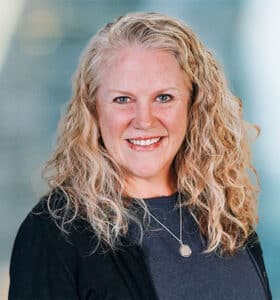 A person with long, curly blonde hair smiles at the camera. They are wearing a dark sweater and a necklace with a round pendant. The background is softly blurred, giving a professional ambiance.