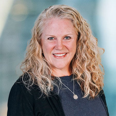 A person with long, curly blonde hair smiles at the camera. They are wearing a dark sweater and a necklace with a round pendant. The background is softly blurred, giving a professional ambiance.