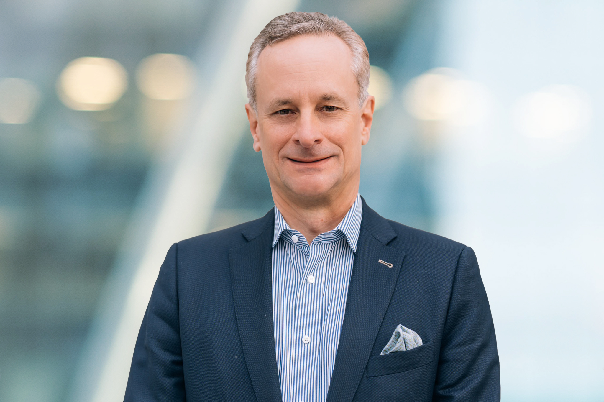 A middle-aged man in a dark suit jacket and striped shirt stands smiling in front of a blurry modern glass building background. He has short gray hair and a pocket square in his jacket.