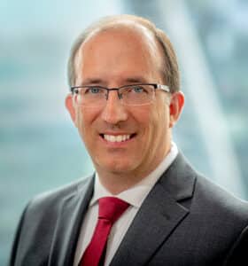 A smiling man in a gray suit, white shirt, and red tie, with short hair and glasses, stands in front of a blurred background of an office building.