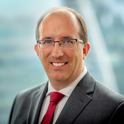 A smiling man in a gray suit, white shirt, and red tie, with short hair and glasses, stands in front of a blurred background of an office building.