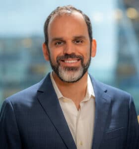 A man in a navy suit and white shirt smiles warmly. He has a beard and stands in front of a large window with a blurred cityscape in the background.