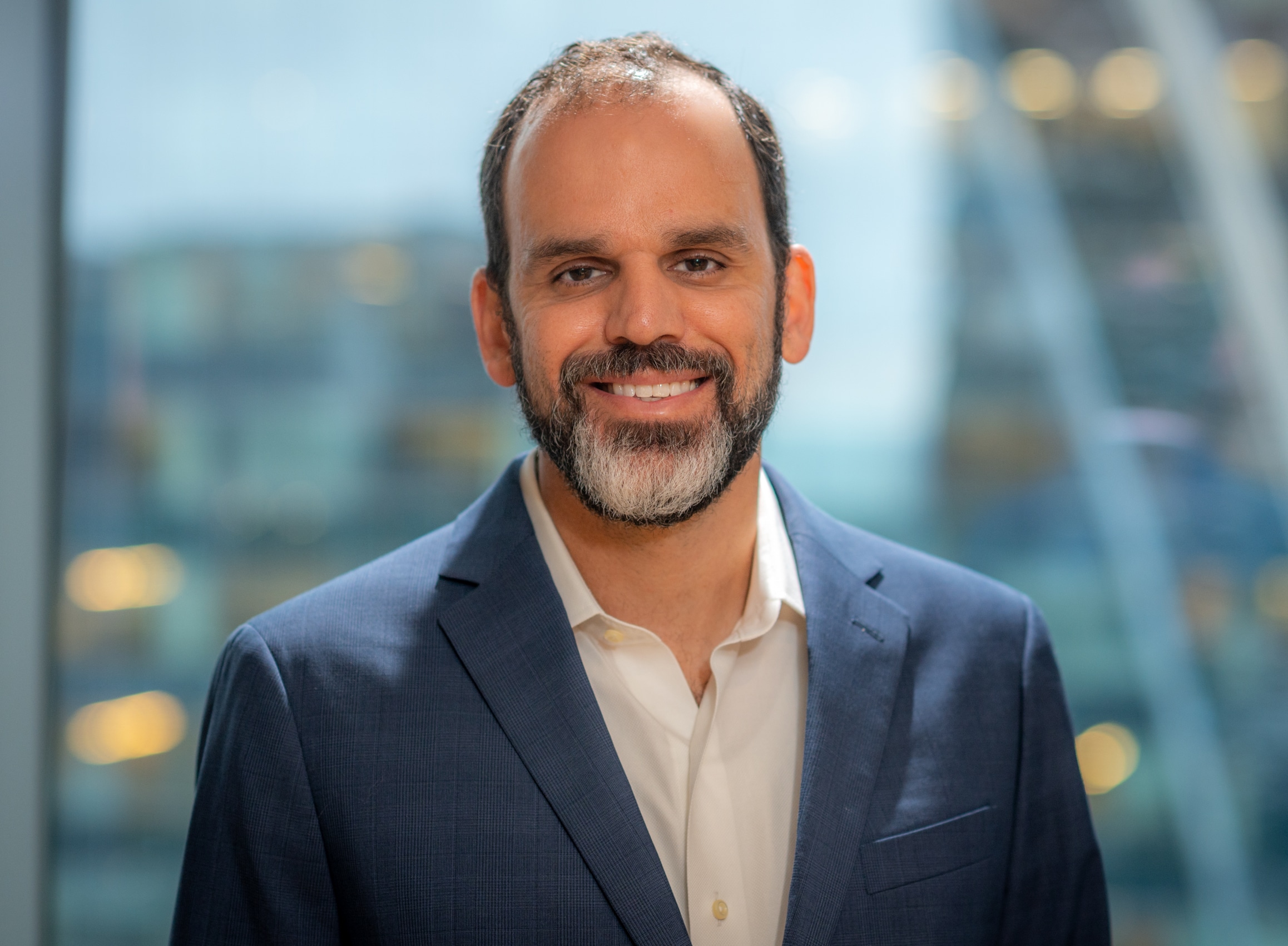 A man in a navy suit and white shirt smiles warmly. He has a beard and stands in front of a large window with a blurred cityscape in the background.