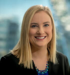 A woman with blonde hair and a black blazer smiles at the camera. She is wearing a blue necklace. The background is softly blurred, showing an indoor setting and possibly a window.