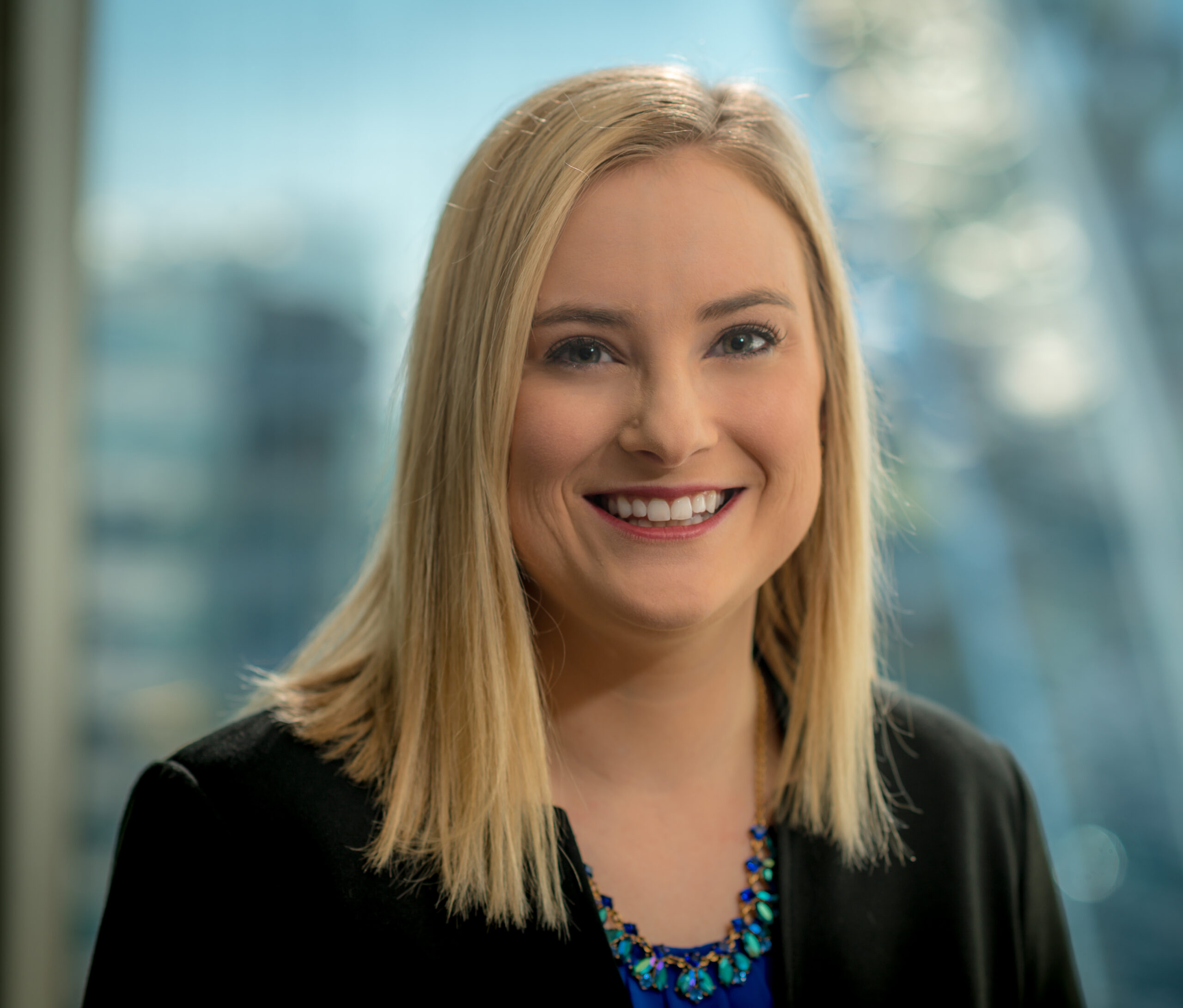A woman with blonde hair and a black blazer smiles at the camera. She is wearing a blue necklace. The background is softly blurred, showing an indoor setting and possibly a window.