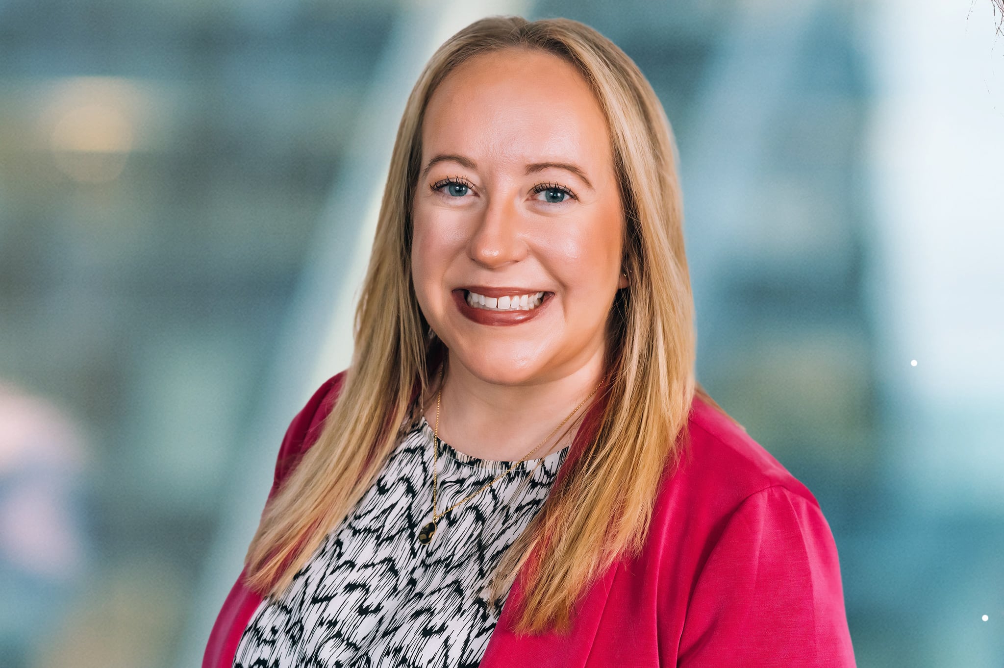 A person with long blonde hair is smiling, wearing a red blazer over a patterned blouse. The background is softly blurred, giving a professional portrait feel.