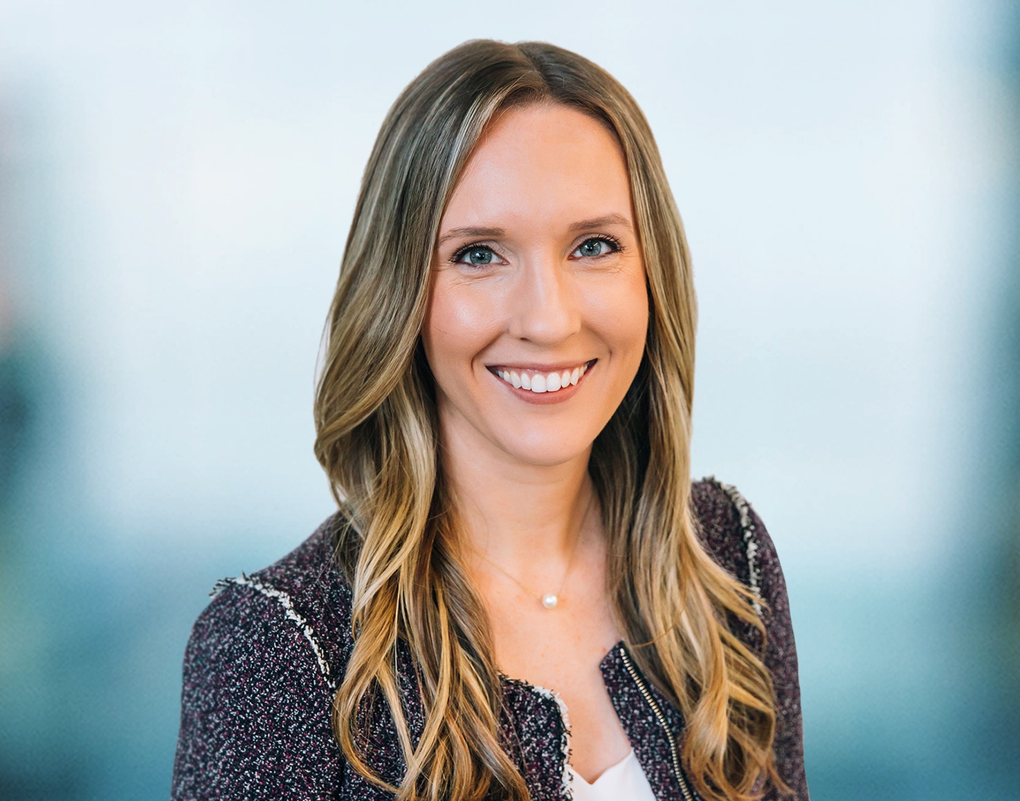 A woman with long, wavy blonde hair smiles at the camera. She is wearing a dark textured blazer over a light top. The background is blurred, giving the image a professional and friendly appearance.