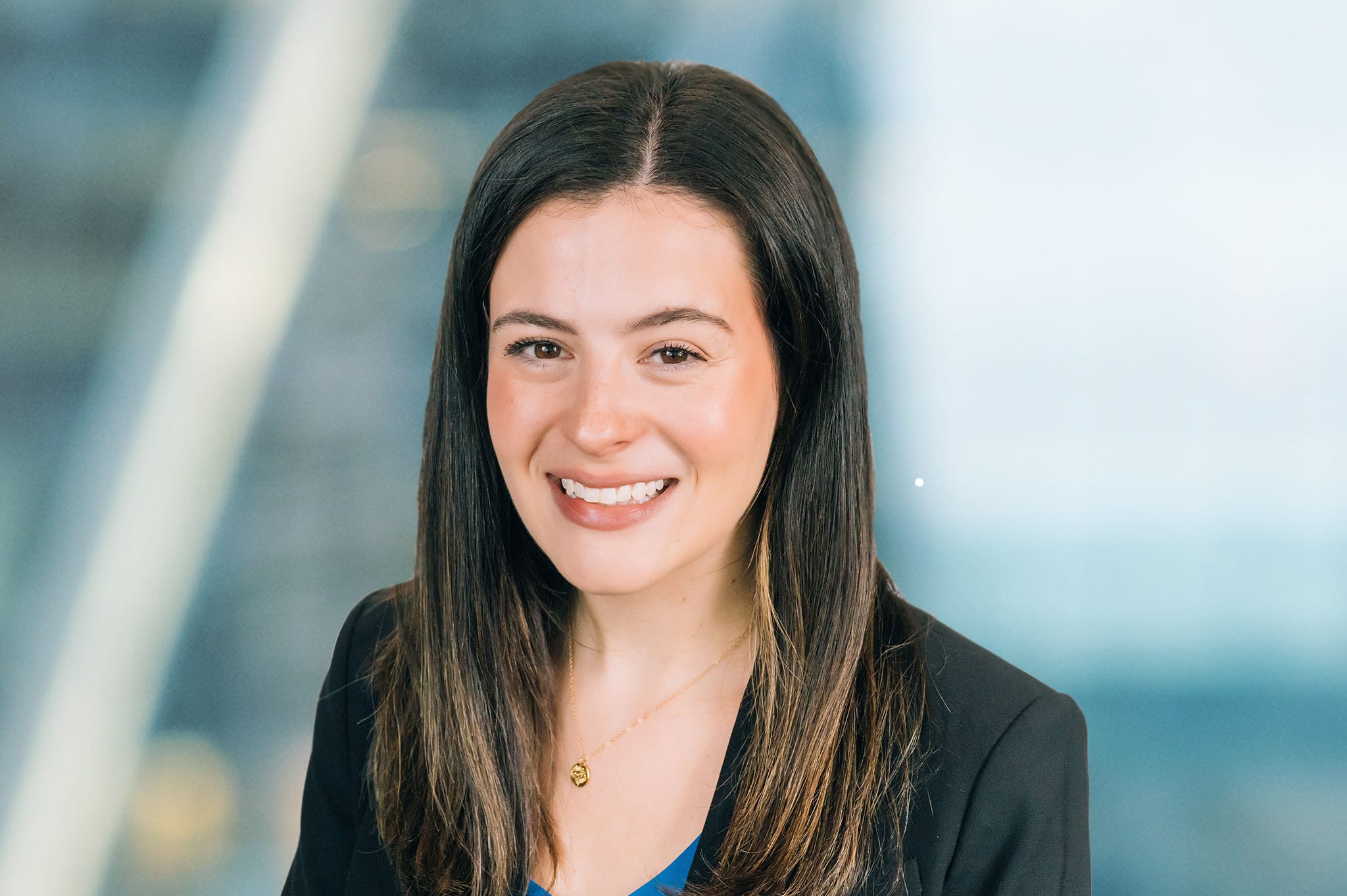 A woman with long dark hair smiles at the camera. She is wearing a dark blazer and a blue top. The background is softly blurred, suggesting an indoor setting with natural light.
