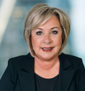A woman with short, light blond hair smiles at the camera. She is wearing a black blazer and a black top. The background is a blurred, light-filled interior setting.