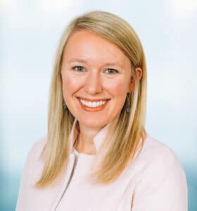 Smiling person with long blonde hair wearing a pink sweater, standing against a soft blue background.