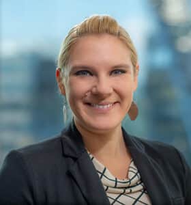 A person with short blond hair smiles while wearing a black blazer and patterned blouse. The background features a blurred view of modern glass buildings.
