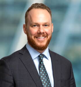 A man with a beard is smiling and wearing a dark suit, light blue shirt, and patterned tie. The background is blurred with a blue and green gradient.