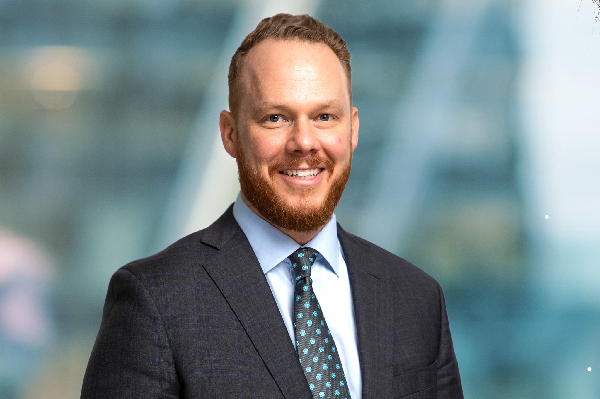 A man with a beard is smiling and wearing a dark suit, light blue shirt, and patterned tie. The background is blurred with a blue and green gradient.