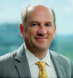 A man in a gray suit and yellow polka dot tie is smiling at the camera. The background is a blurred view of windows and greenery, suggesting an office setting.