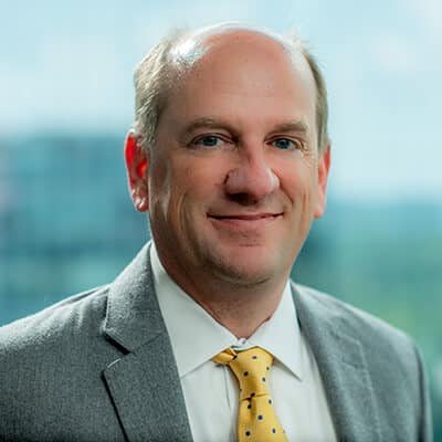 A man in a gray suit and yellow polka dot tie is smiling at the camera. The background is a blurred view of windows and greenery, suggesting an office setting.