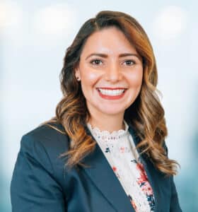A woman with wavy brown hair wearing a navy blazer and a floral blouse smiles at the camera. The background is softly blurred, giving a professional and friendly appearance.