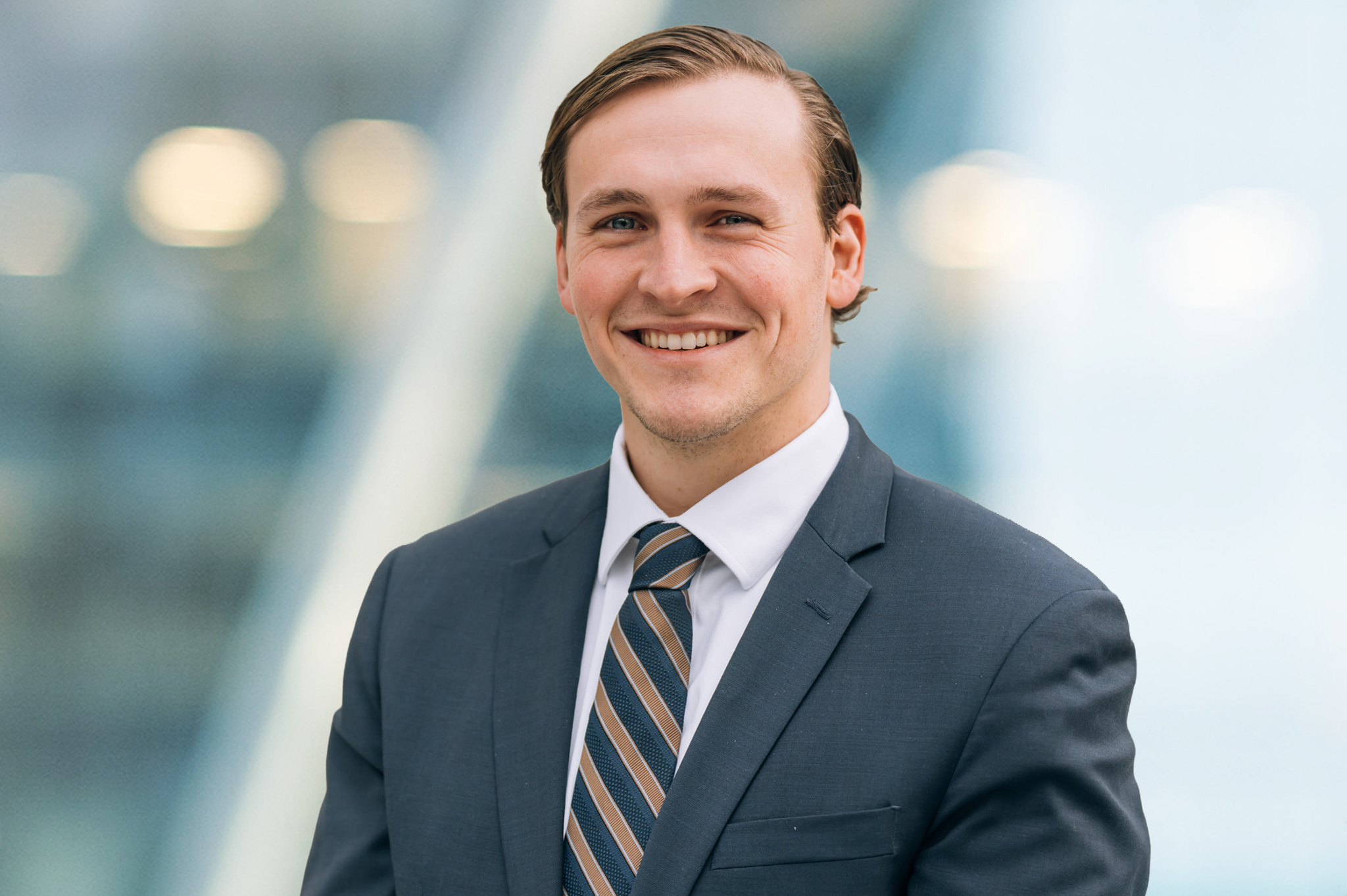 A young man in a dark suit, striped tie, and white shirt smiles at the camera. The background is blurred with soft lighting and modern glass architecture.