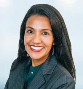 A person with long dark hair and a bright smile, wearing a dark pinstripe blazer and green top, is looking at the camera against a light blue background.