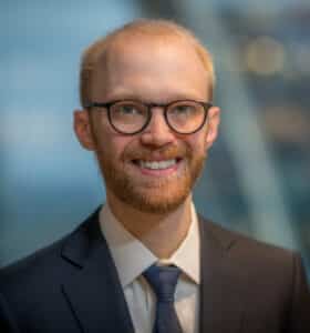 A smiling person with glasses and a beard wears a suit and tie. The background is softly blurred, suggesting an indoor setting with modern architecture elements.