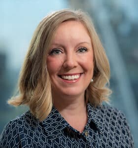 A smiling person with shoulder-length blond hair is wearing a patterned dark shirt. The background is blurred with soft blue and white tones, suggesting an indoor setting with natural light.