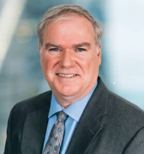 A middle-aged man in a dark gray suit and blue dress shirt smiles at the camera. He has short gray hair and is wearing a patterned blue tie. The background is blurred and appears to be an indoor setting.