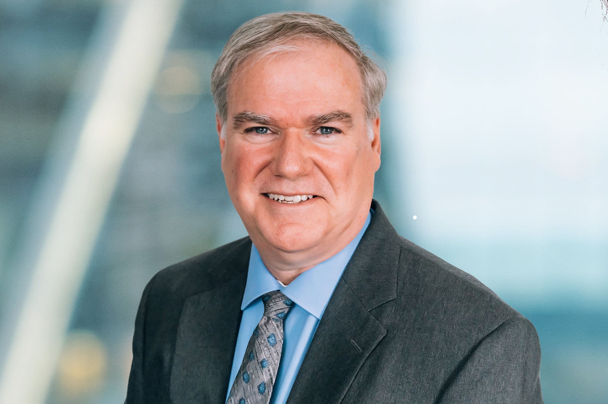 A middle-aged man in a dark gray suit and blue dress shirt smiles at the camera. He has short gray hair and is wearing a patterned blue tie. The background is blurred and appears to be an indoor setting.