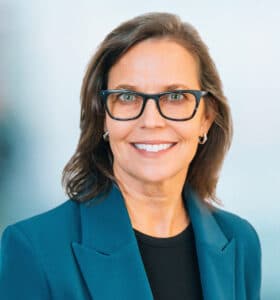 A woman with shoulder-length brown hair and glasses smiles at the camera. She is wearing a teal blazer over a black top. The background is a blurred light blue and gray gradient.