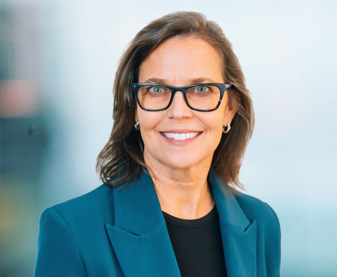 A woman with shoulder-length brown hair and glasses smiles at the camera. She is wearing a teal blazer over a black top. The background is a blurred light blue and gray gradient.