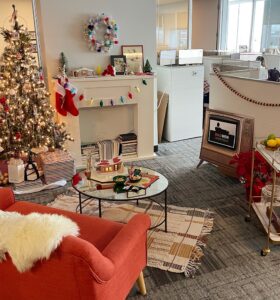 An office space decorated for the holidays with a Christmas tree, stockings on a faux fireplace, a red sofa with a fur throw, and a vintage TV. A bar cart is adorned with festive decorations, enhancing the cozy, festive atmosphere.