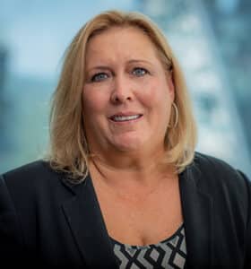 A woman with blonde hair is wearing a black blazer and a patterned top. She is standing indoors with a blurred background of windows and greenery.