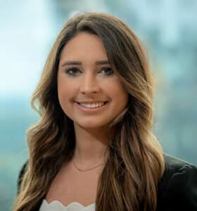 A woman with long brown hair is smiling at the camera. She is wearing a black blazer and a white top. The background is blurred with hints of green and blue, suggesting an outdoor or window view.