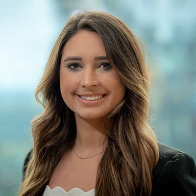 A woman with long brown hair is smiling at the camera. She is wearing a black blazer and a white top. The background is blurred with hints of green and blue, suggesting an outdoor or window view.