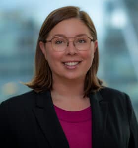 A person with shoulder-length brown hair, wearing glasses, a black blazer, and a magenta top, smiles in front of a softly blurred urban background.