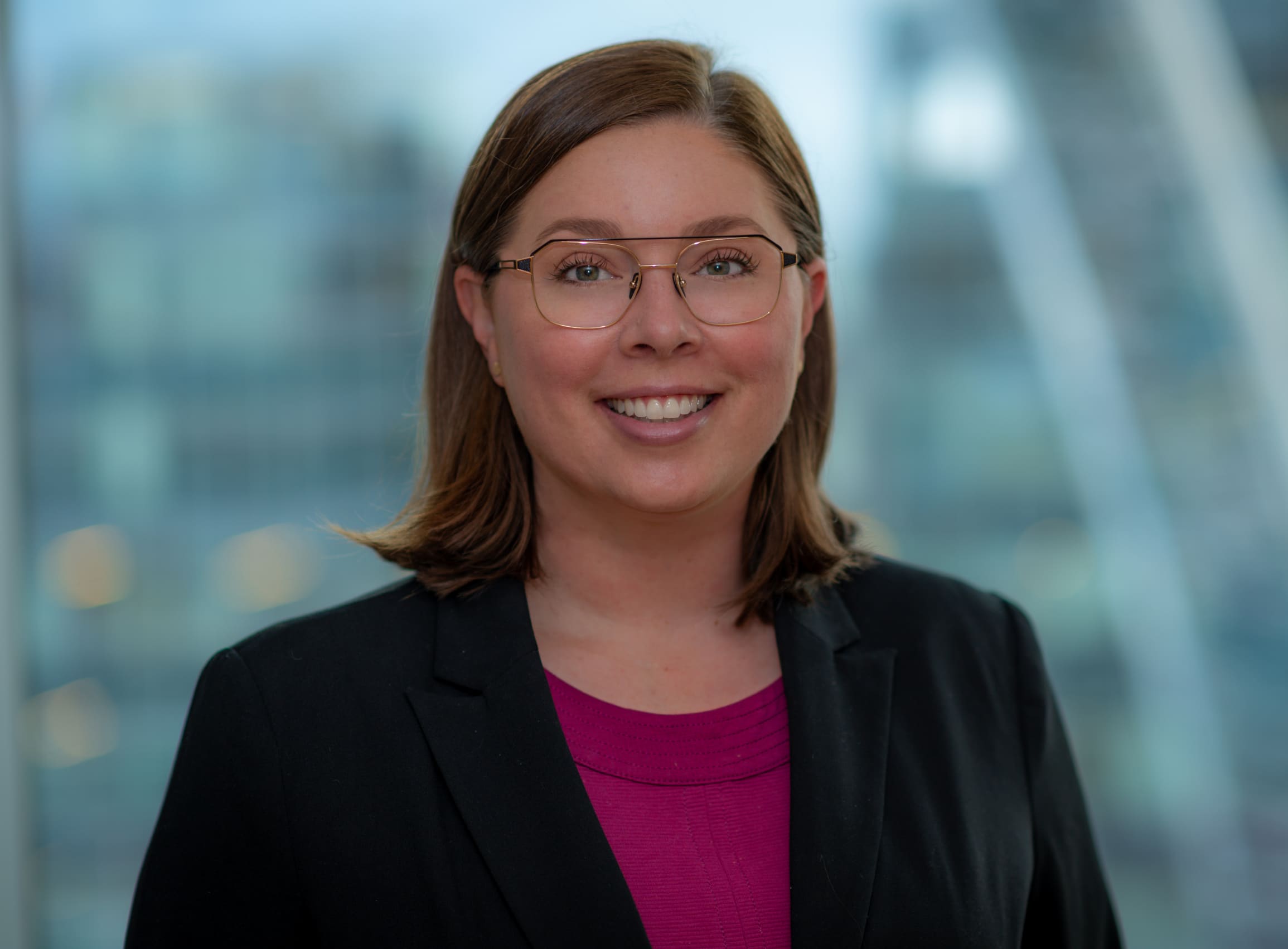 A person with shoulder-length brown hair, wearing glasses, a black blazer, and a magenta top, smiles in front of a softly blurred urban background.