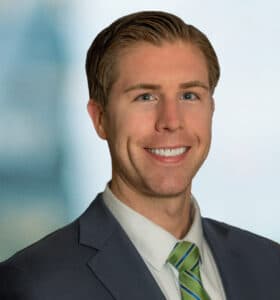 A smiling man in a suit with a green and white striped tie stands against a blurred blue background.
