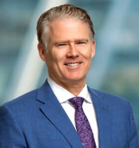 A smiling man in a blue suit and purple tie stands in front of a blurred background, possibly a cityscape.