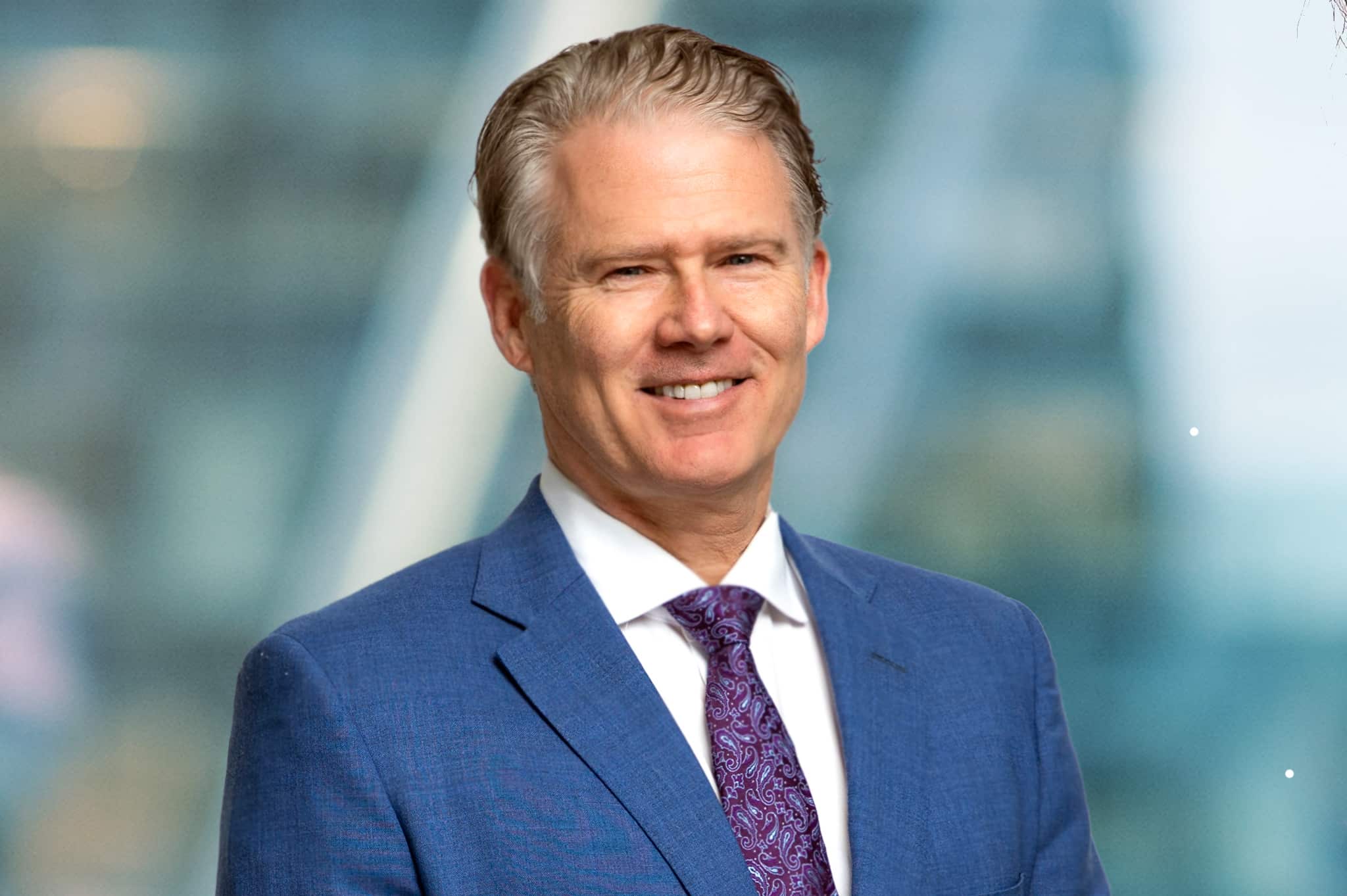 A smiling man in a blue suit and purple tie stands in front of a blurred background, possibly a cityscape.