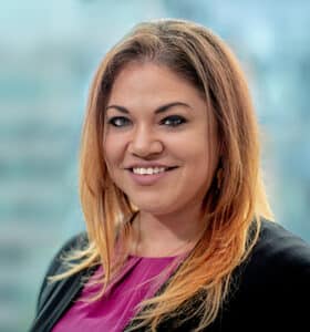 A woman with long, light brown hair and a friendly smile. She is wearing a magenta top and black blazer. The background is softly blurred, suggesting an office setting.