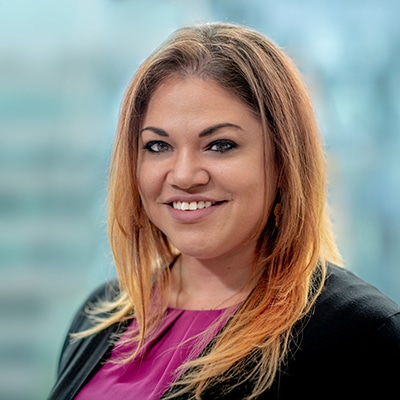 A woman with long, light brown hair and a friendly smile. She is wearing a magenta top and black blazer. The background is softly blurred, suggesting an office setting.