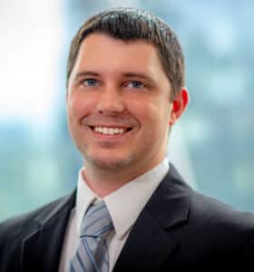 A man in a suit and tie smiles while standing against a blurred background of windows and greenery.