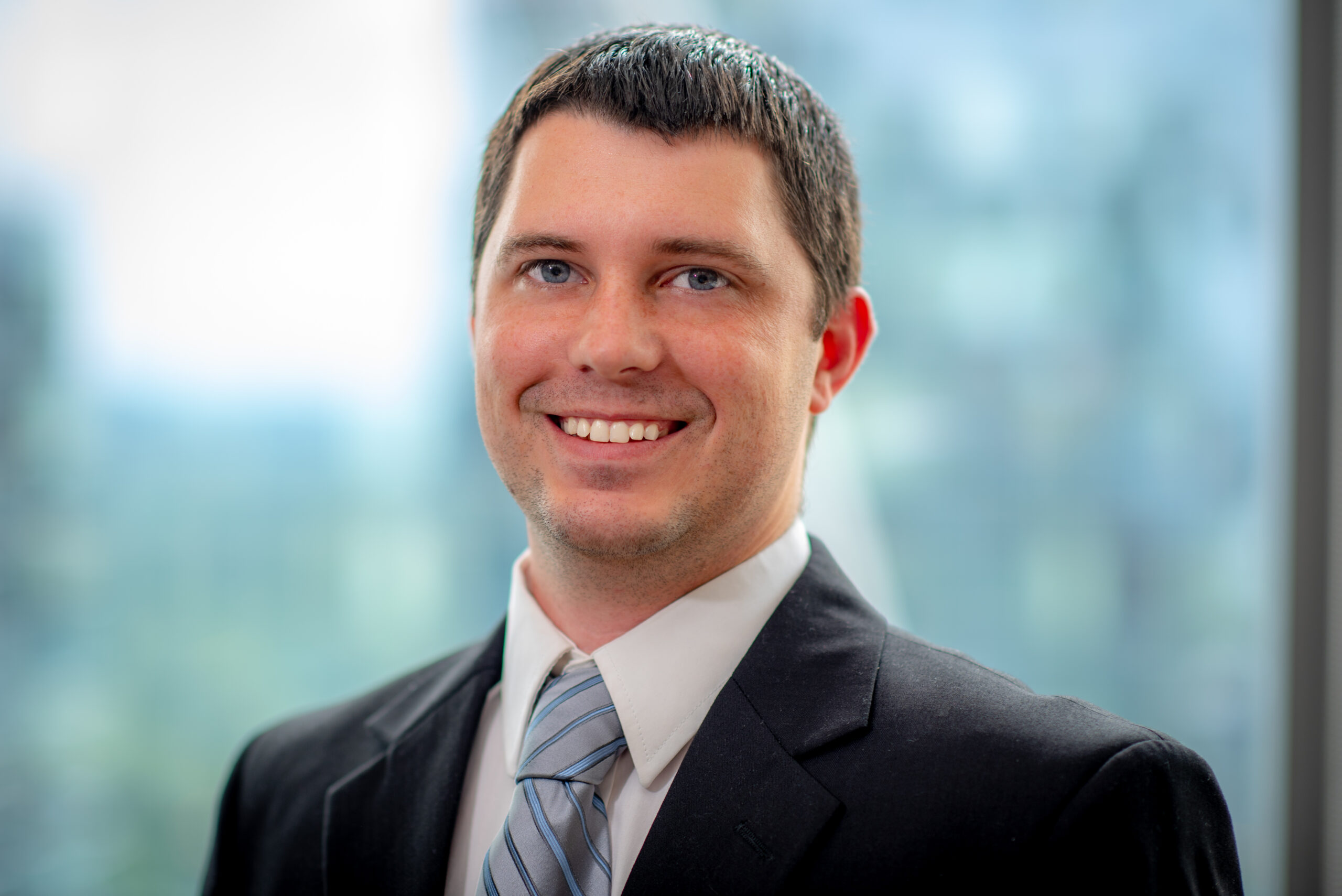 A man in a suit and tie smiles while standing against a blurred background of windows and greenery.