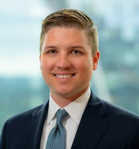 A person in a suit and tie smiles at the camera. The background is blurred, suggesting an indoor setting with a window view.