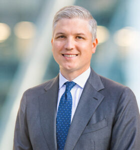 A man with short gray hair in a gray suit, white shirt, and blue polka dot tie smiles while standing in front of a blurred, modern city background.