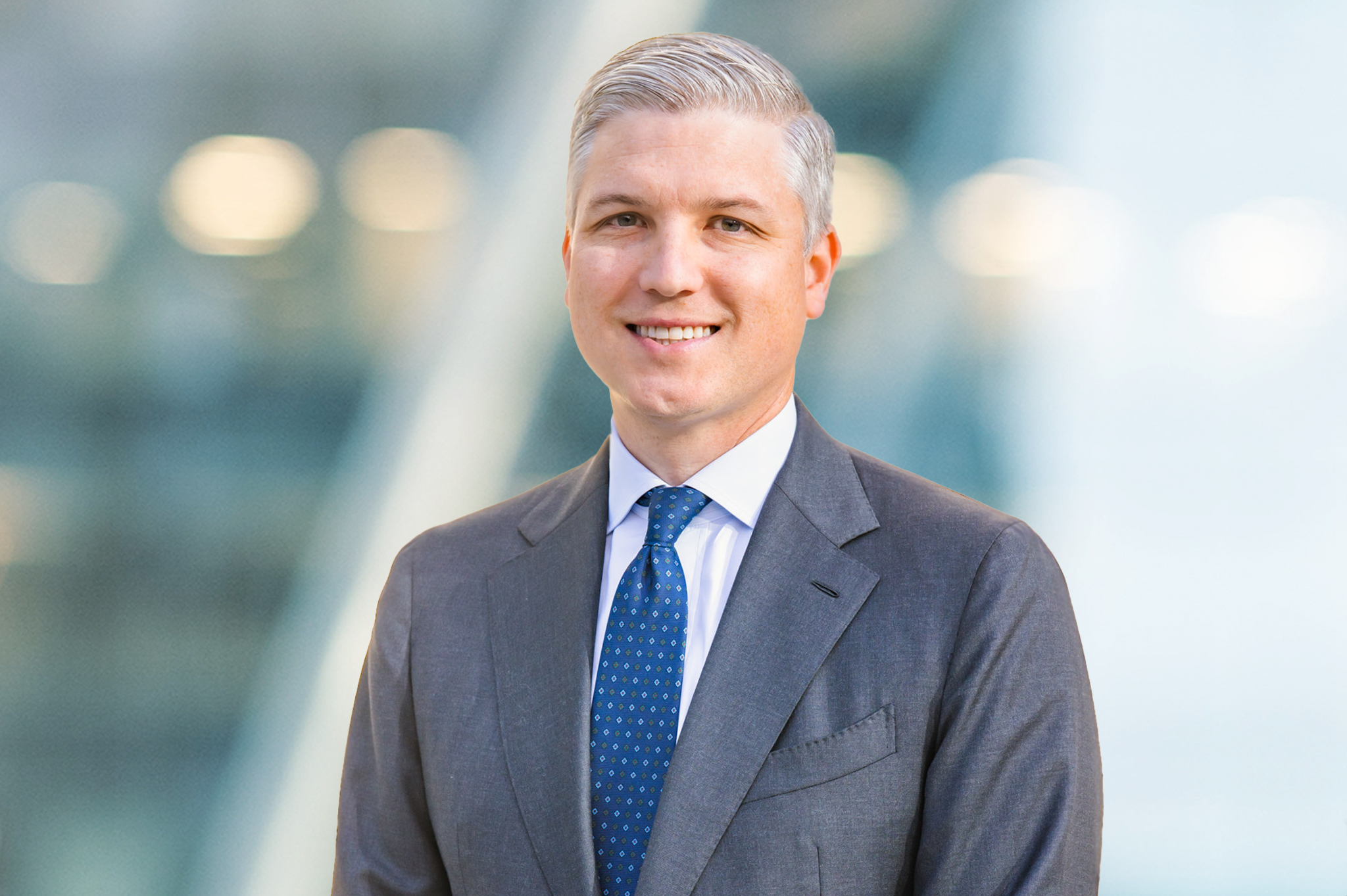 A man with short gray hair in a gray suit, white shirt, and blue polka dot tie smiles while standing in front of a blurred, modern city background.