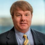 A person with light brown hair wearing a suit, light blue shirt, and yellow striped tie, standing in front of a glass window. The background is blurred.
