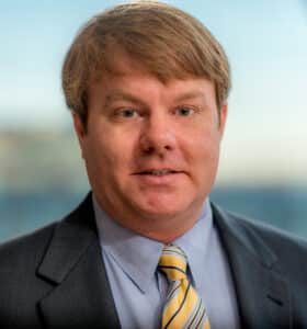 A person with light brown hair wearing a suit, light blue shirt, and yellow striped tie, standing in front of a glass window. The background is blurred.