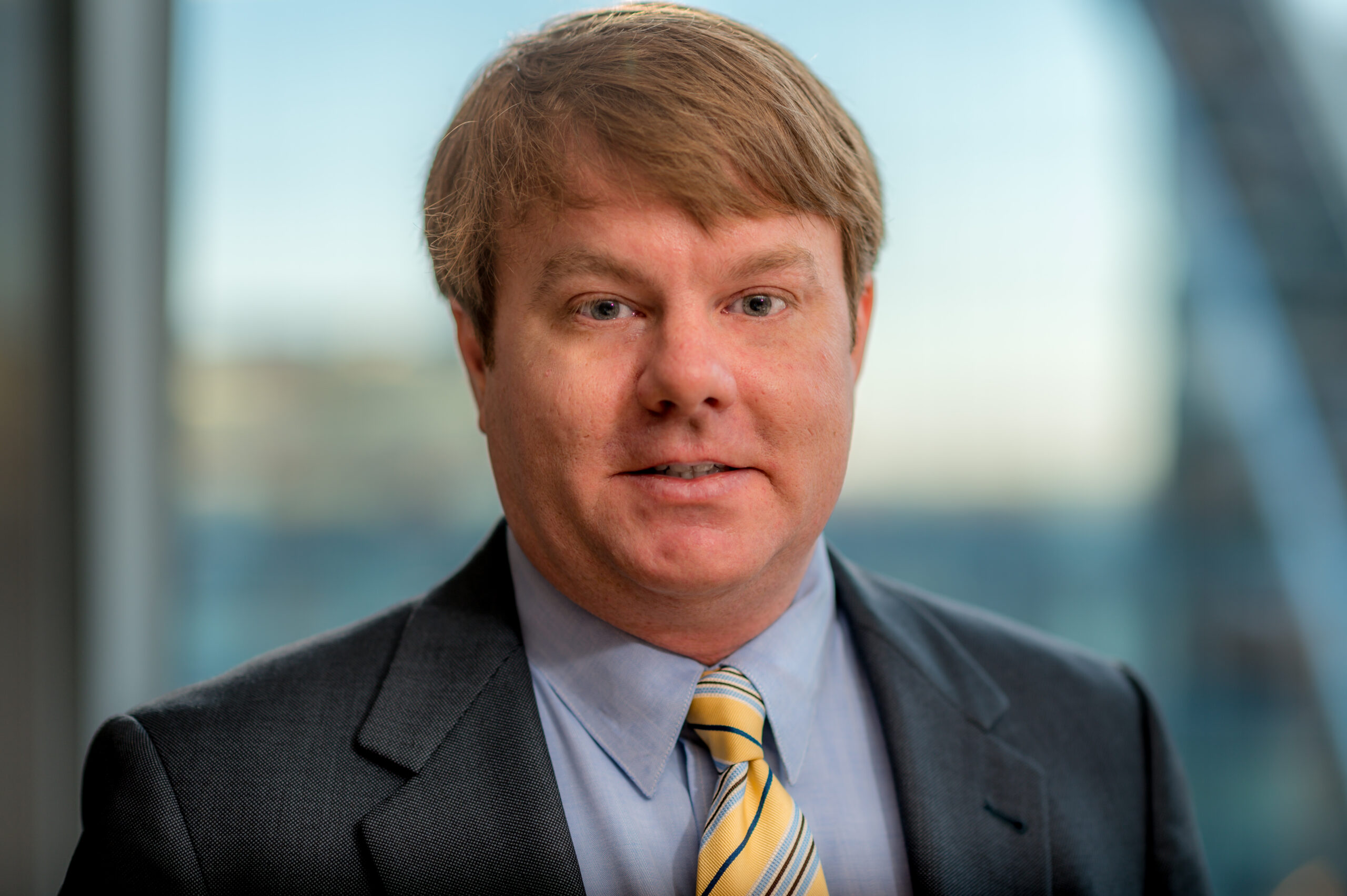 A person with light brown hair wearing a suit, light blue shirt, and yellow striped tie, standing in front of a glass window. The background is blurred.