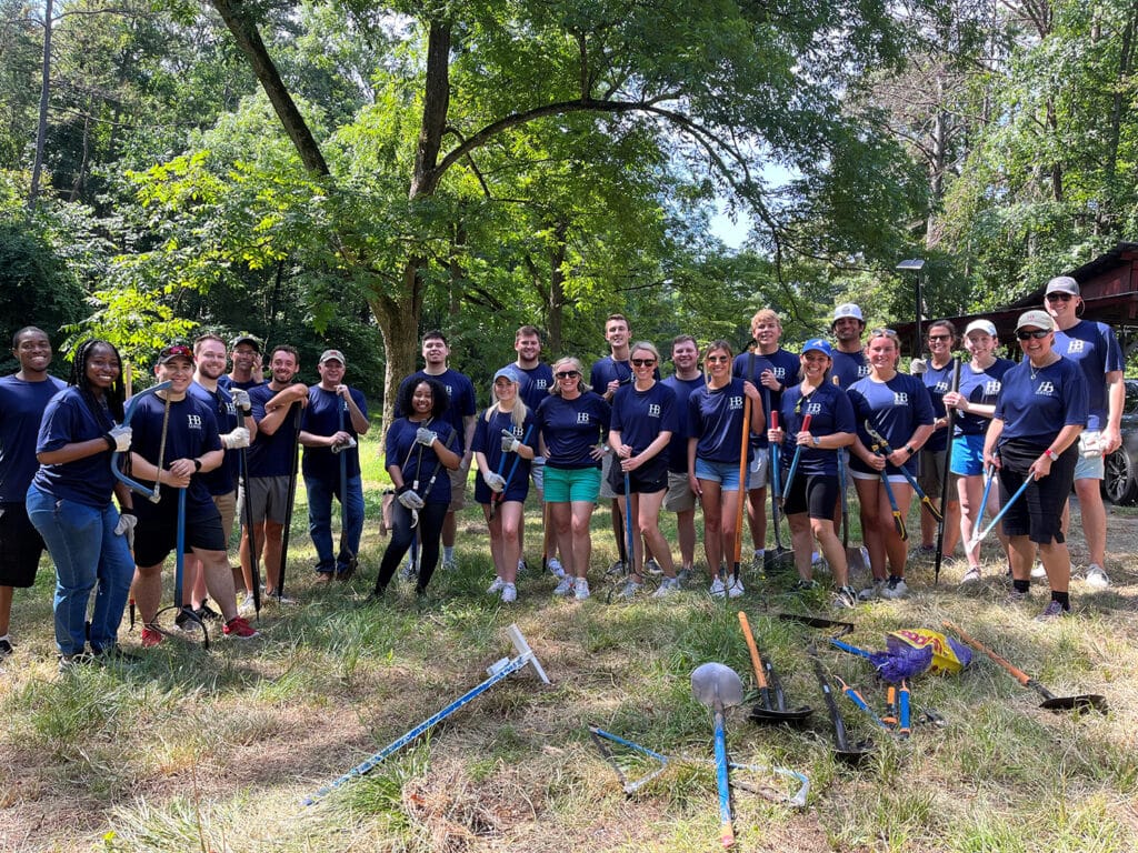 A diverse group of people in matching blue shirts stand together outdoors in a wooded area, holding gardening tools and smiling. They appear to be participating in a community cleanup or environmental activity.